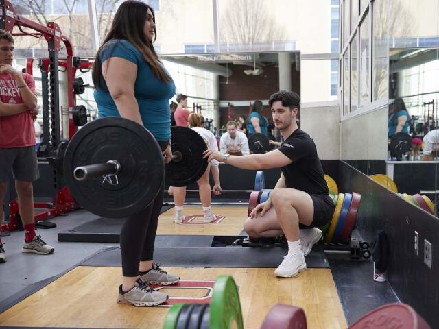 Personal trainer coaches a participant performing a barbell lift in the RPAC Fitness Suite.